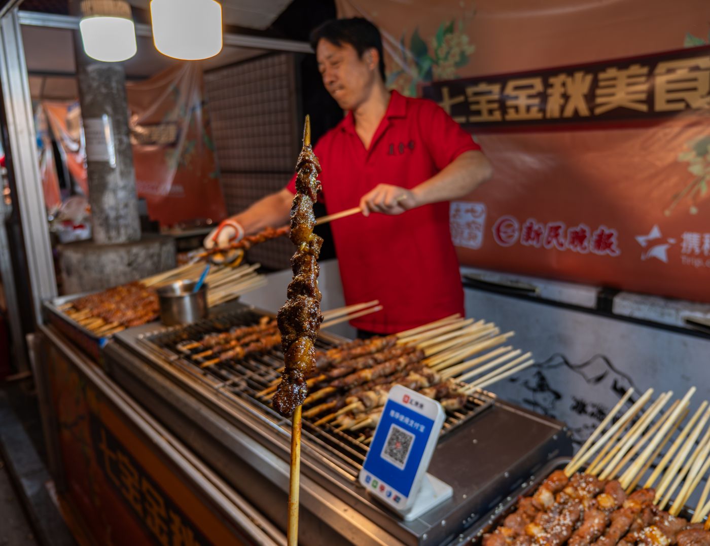 国内夜市摊主在摊前烤串，背景是中文招牌与扫码点单牌